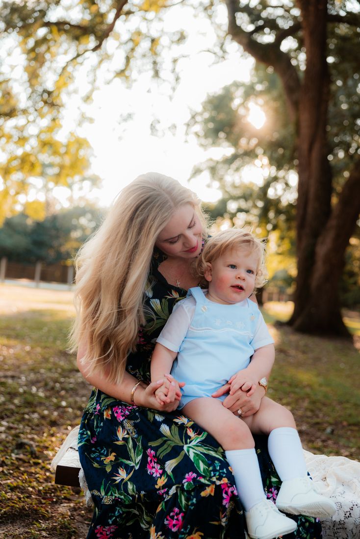 A mother in a floral dress sitting with her little boy on her lap on a bench under the trees