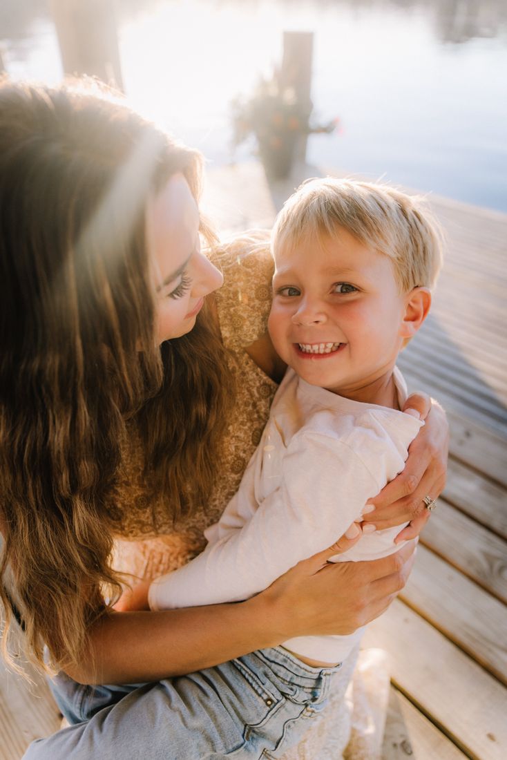 A mother looking down at her son as she holds him on the dock, the water bright behind them