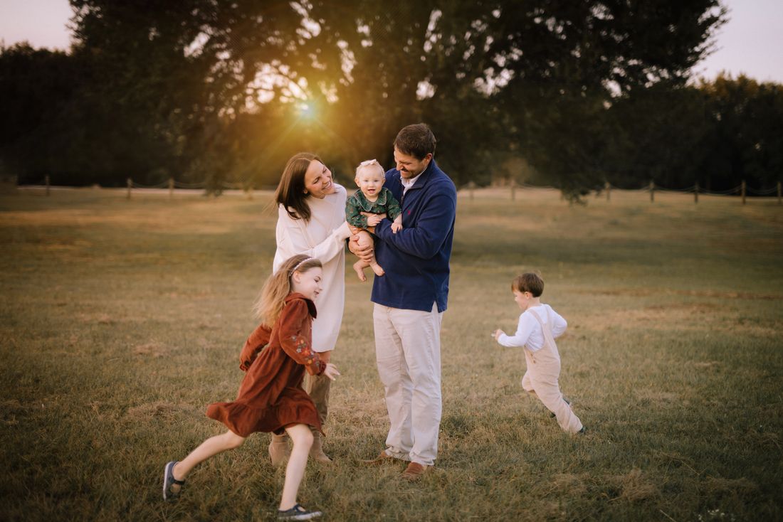 A family playing together in a field at sunset, the older two children running circles around their parents