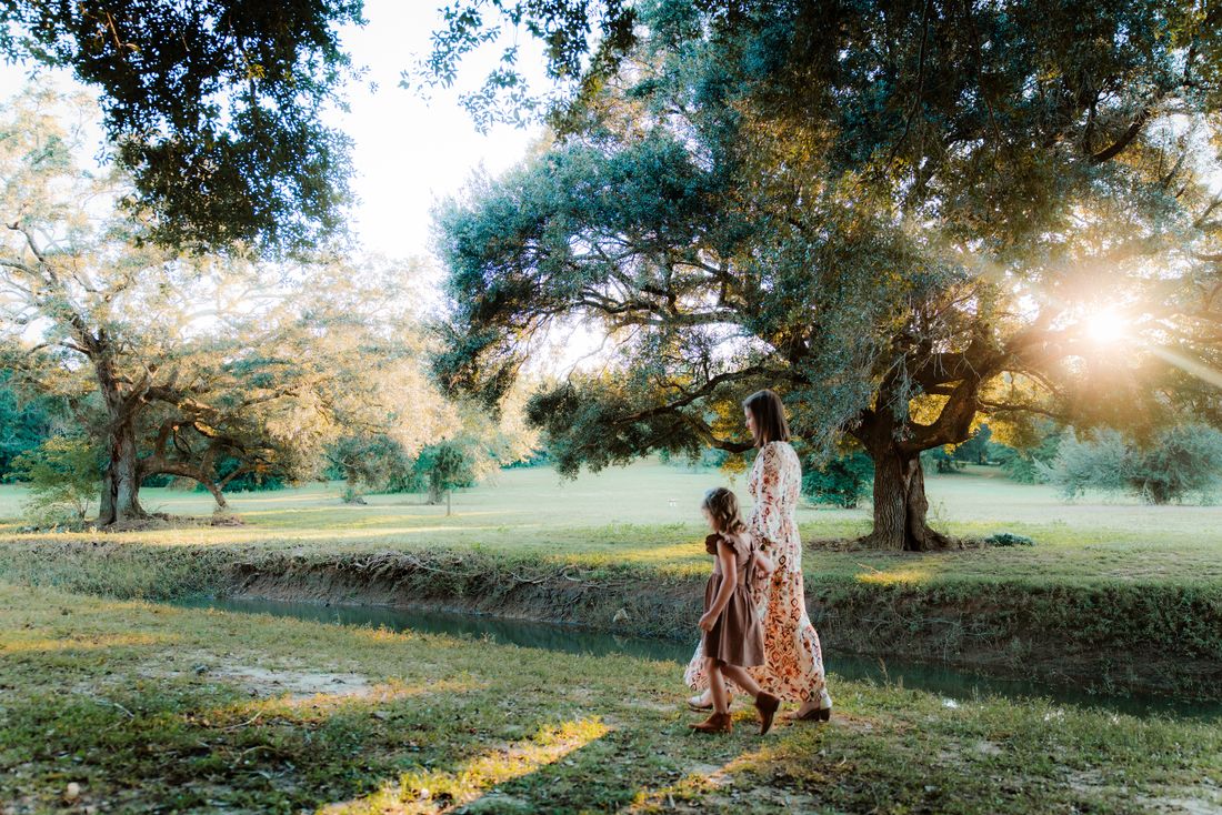 A mother and her little one small in the frame beneath a wide live oak, the sun low behind them