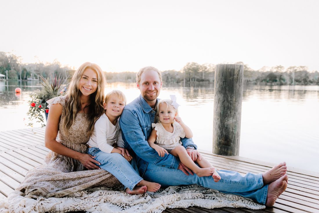 A family of four sitting barefoot on a lace blanket at the end of a dock, the water bright behind them