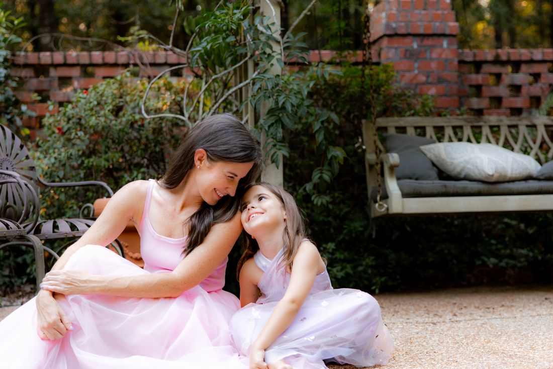 A mother and daughter in pink leaning into each other on a garden patio, smiling at one another