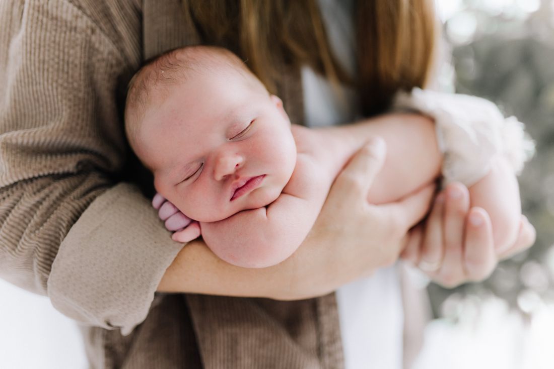 A newborn asleep along his mother's forearm, cheek resting on her hand in soft window light