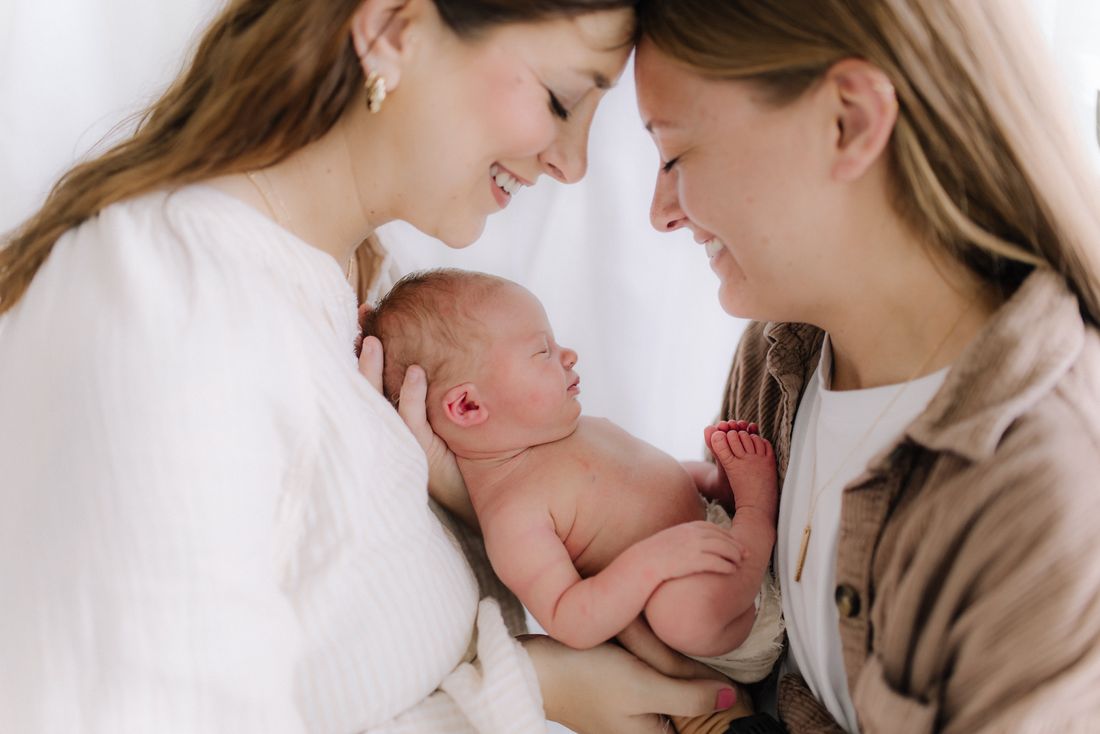 Two mothers forehead to forehead, smiling over the newborn held between them