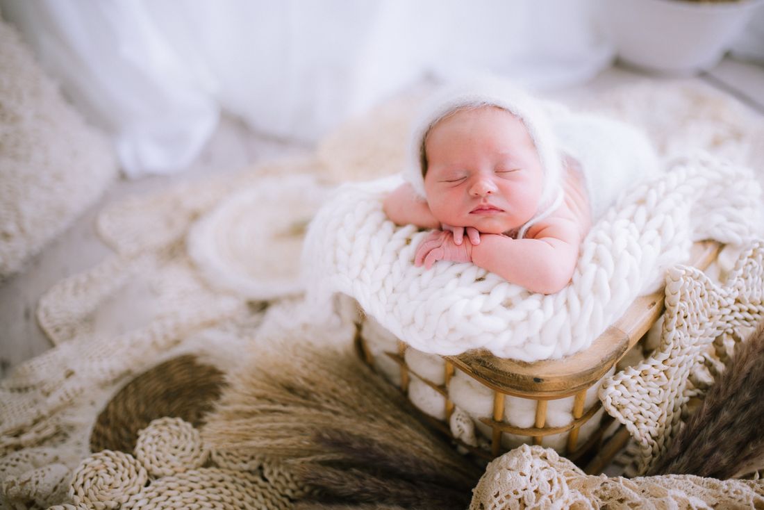 A newborn in a knit bonnet asleep over folded arms in a wooden crate