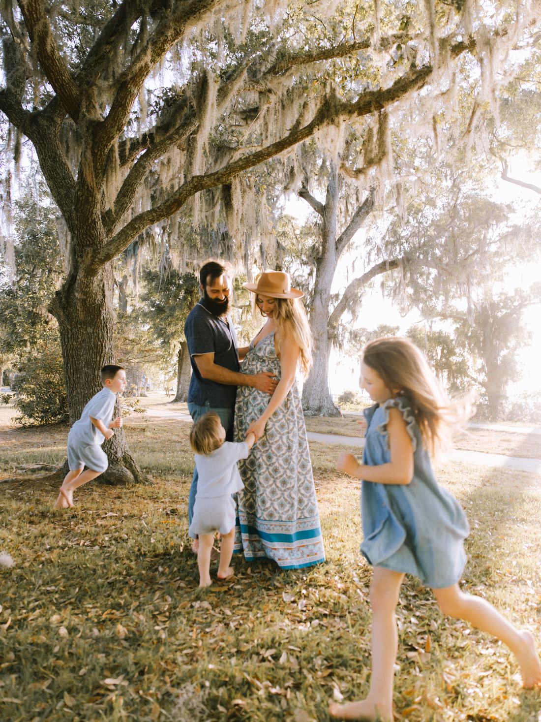 An expecting couple under a moss-draped live oak while their three children run barefoot around them