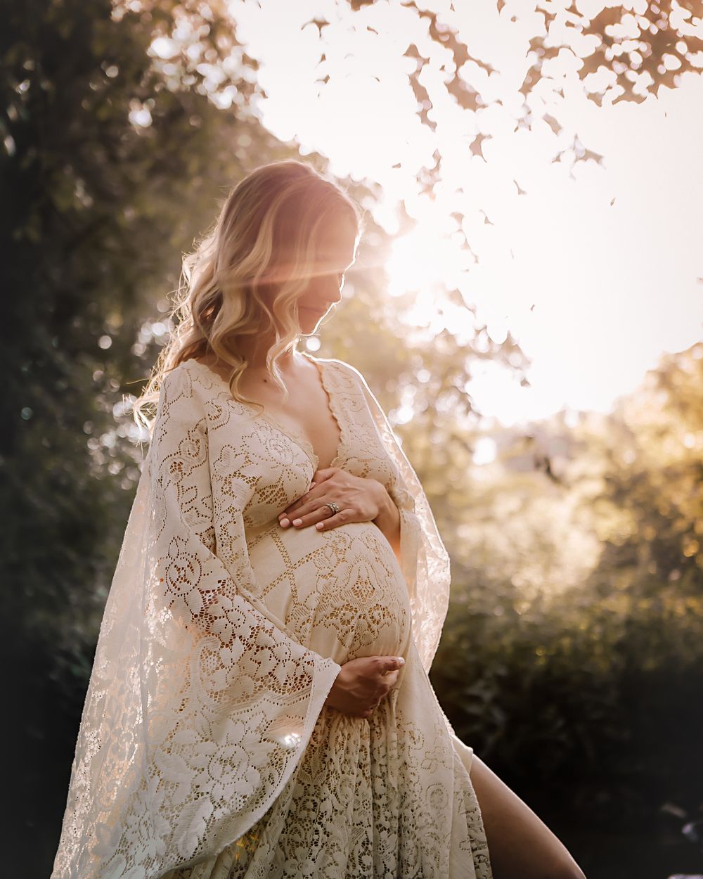 An expecting mother in a flowing lace gown, cradling her belly in backlit golden light by the water