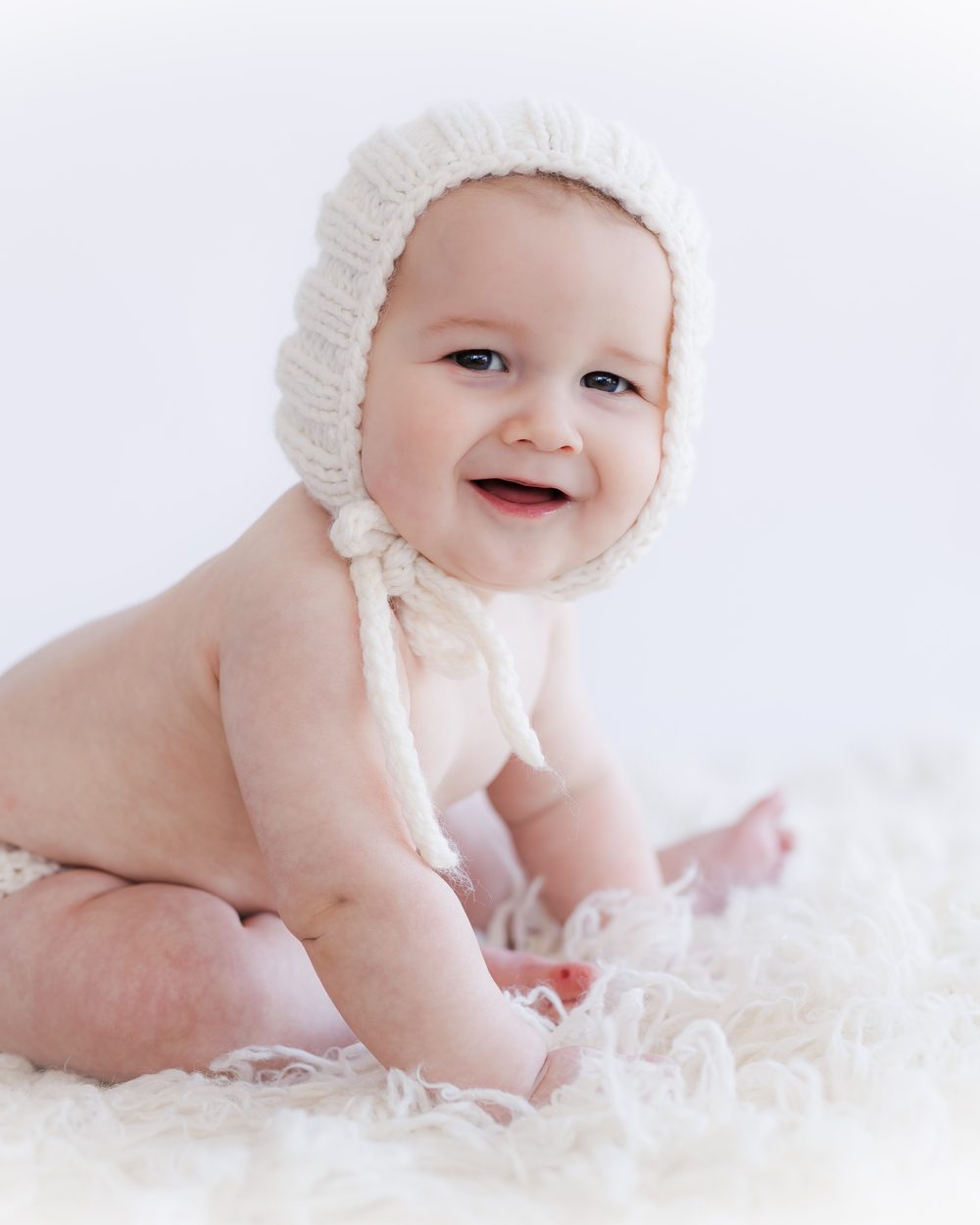 A smiling baby in a cream knit bonnet sitting up on a soft sheepskin in the studio