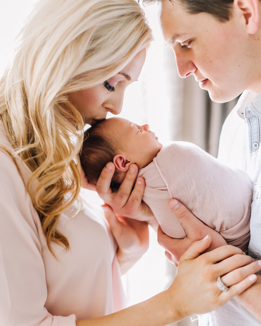 New parents in soft window light, mother kissing her swaddled newborn's head as the father holds them both