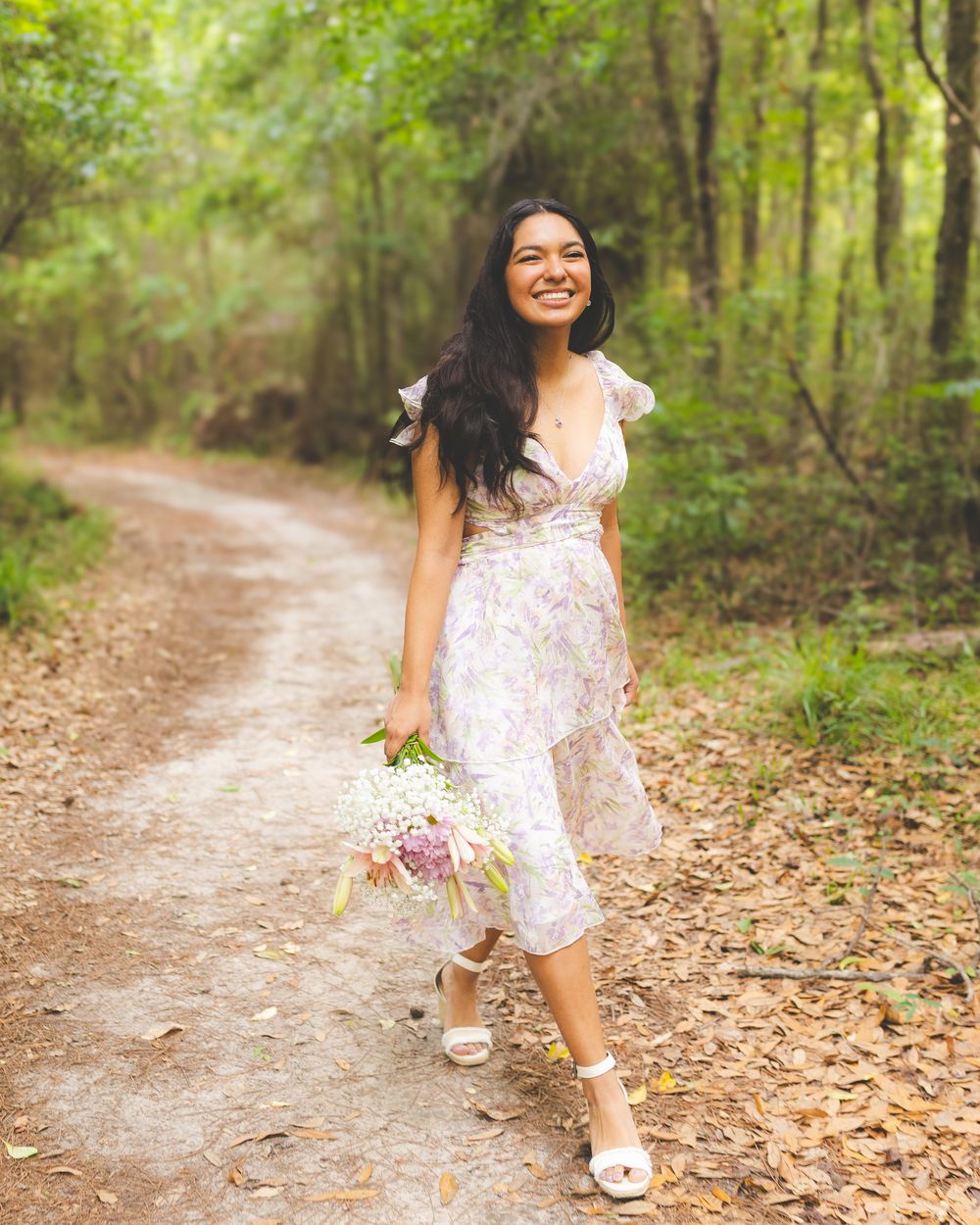 A high school senior walking a wooded path in a floral dress, holding a bouquet and laughing